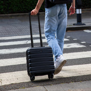 Person with a black backpack walking on a street with trees and a crosswalk in the background.